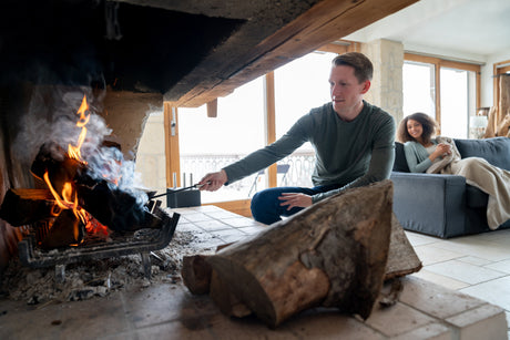 Man tending to a fire in a large indoor fireplace while a woman relaxes on the couch in the background.