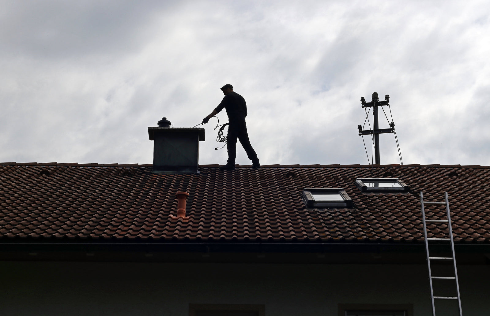 Worker standing on a tiled roof, inspecting or cleaning a chimney under a cloudy sky.