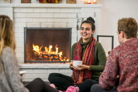 Group of friends sitting by a fireplace, one holding a cup and smiling.