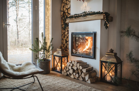 Cozy living room with a lit fireplace, stacked firewood, holiday decor, and large windows overlooking a winter landscape.